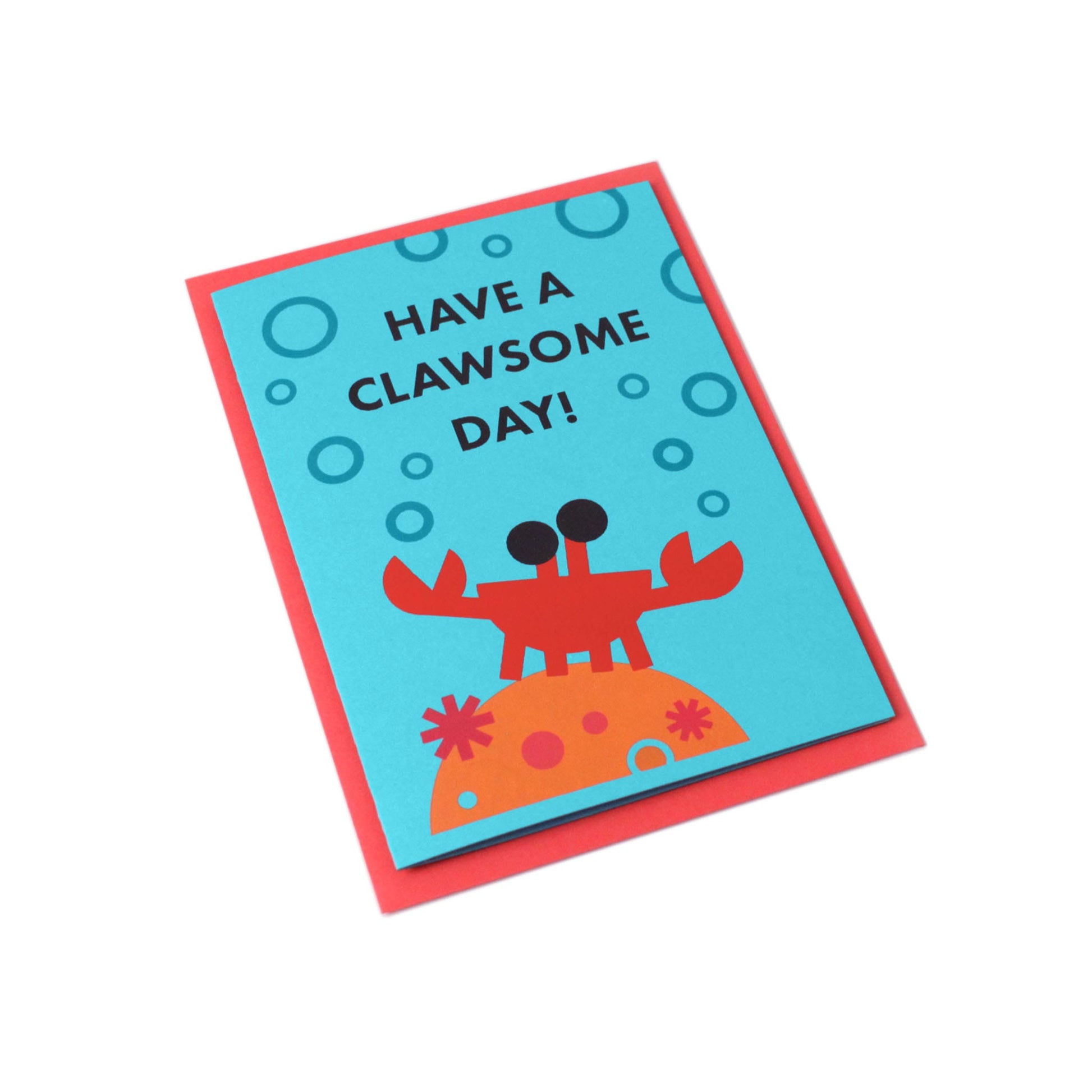 Blue card with text Have a clawsome day! and an illustration of a crab, coral, and bubbles. Red envelope in the background.