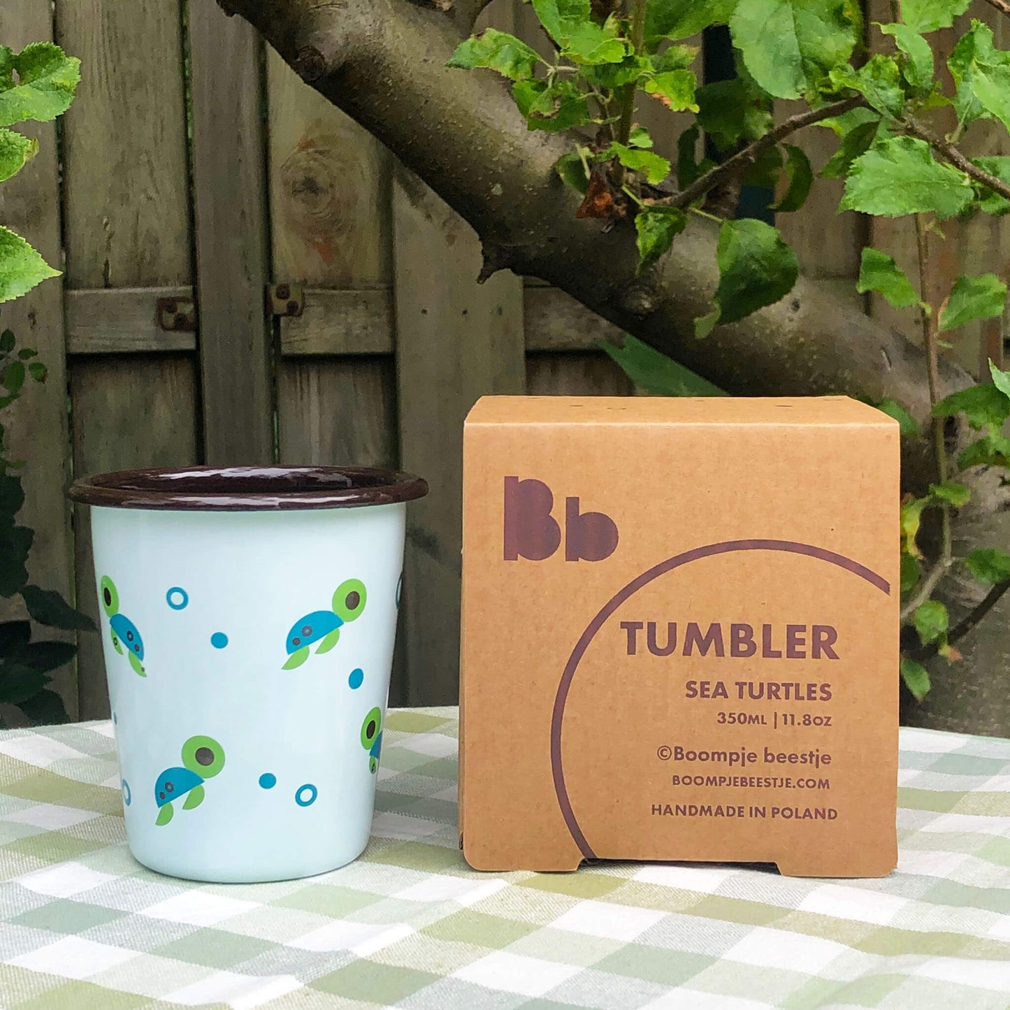 Light blue mug with sea animals illustrations, beside a cardboard box with text Tumbler SEA TURTLES 350ml 11.8oz, placed on a table in a garden.