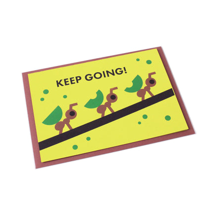 A tilted yellow-green greeting card with the text 'Keep going!' and an illustration of ants carrying leaves on a branch. A brown envelope is visible behind the card.