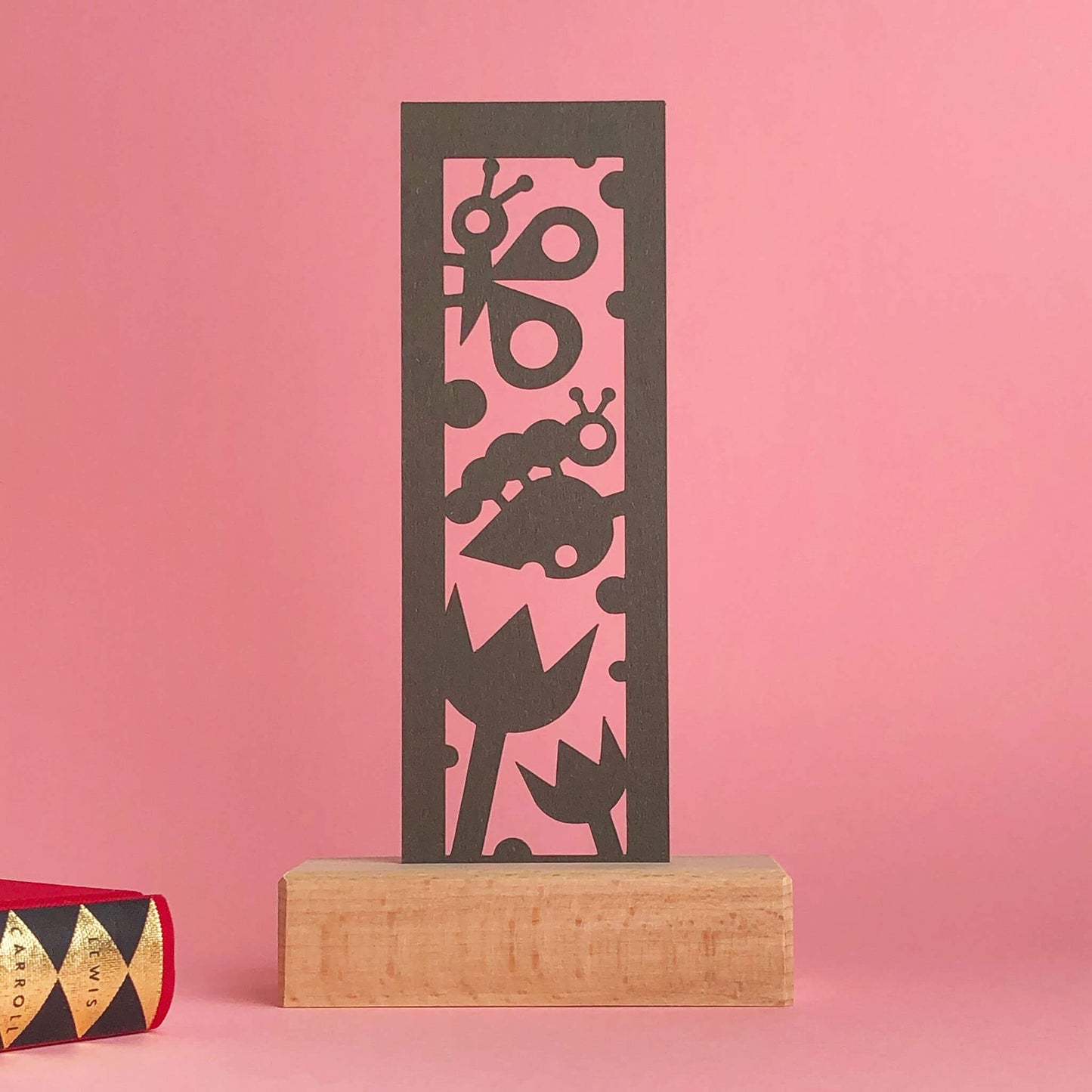 A wooden art display features a brown paper bookmark with a tulip, butterfly and caterpillar cut-out design. A glimpse of a book can be seen beside the bookmark against a pink background.