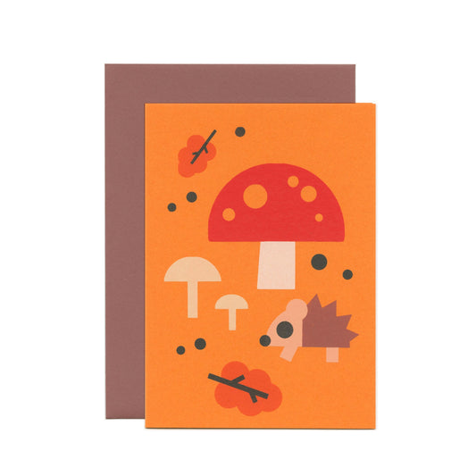 An orange greeting card with an illustration of a hedgehog, mushrooms, leaves and dots. Behind the card is a brown envelope.