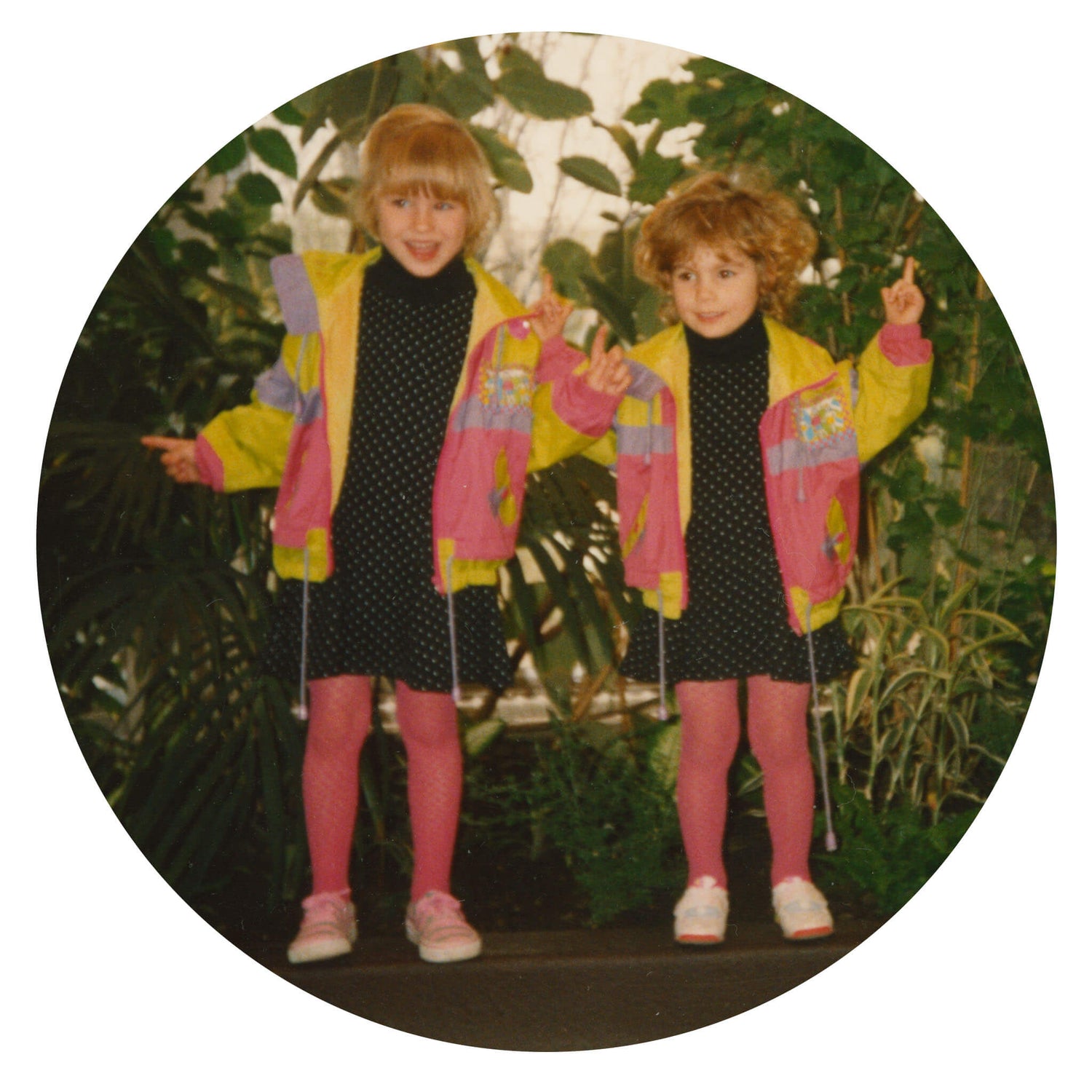 Two young girls, dressed in matching outfits, smiling while lifting their fingers in the air against a background of green plants.