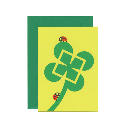 A green greeting card with a four-leaf clover and ladybugs illustrated. There's a dark green envelope behind the card.
