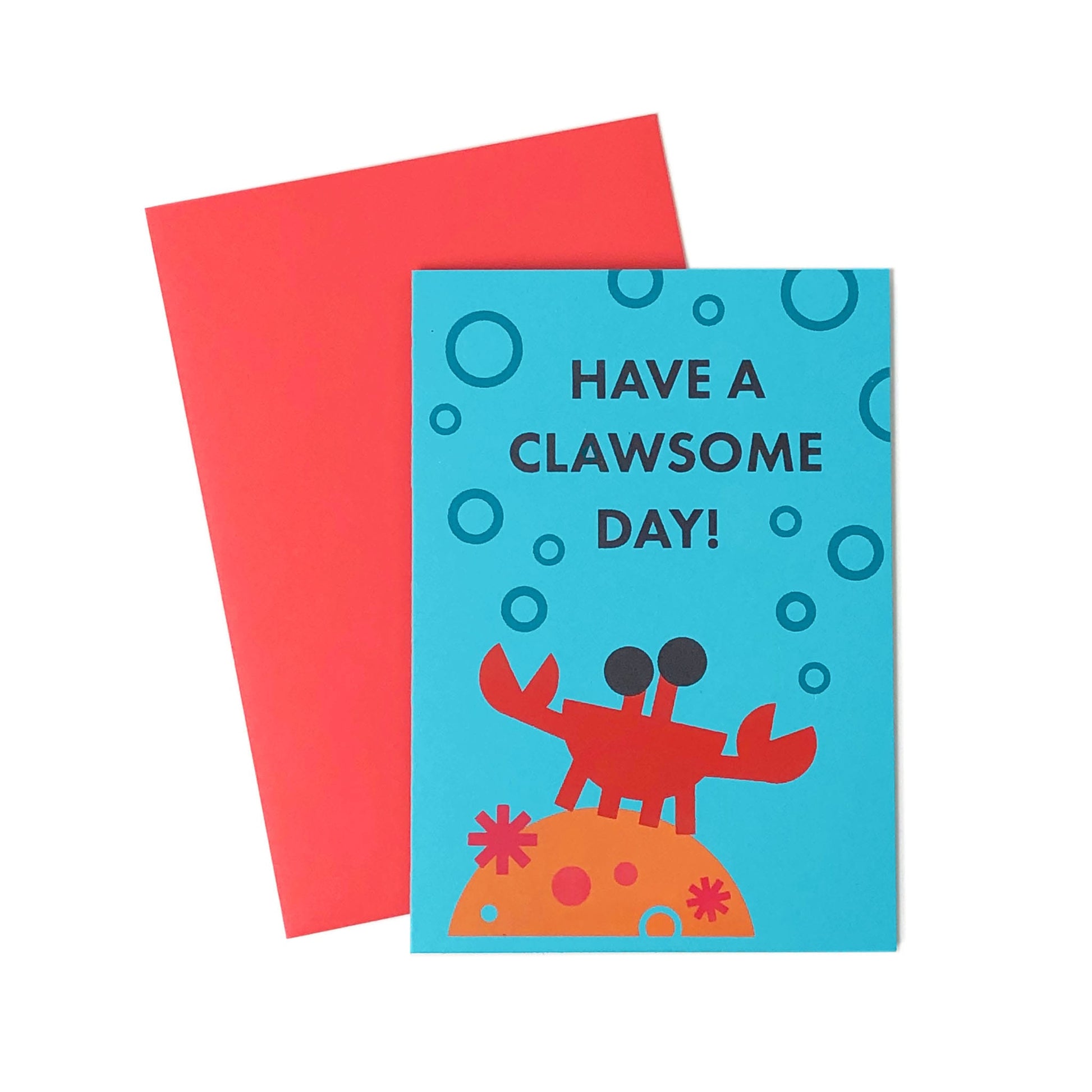 A blue greeting card with an illustration of a crab on coral and bubbles.  On the card is a text that says "Have a clawsome day!". Behind the card is a red-pink envelope.