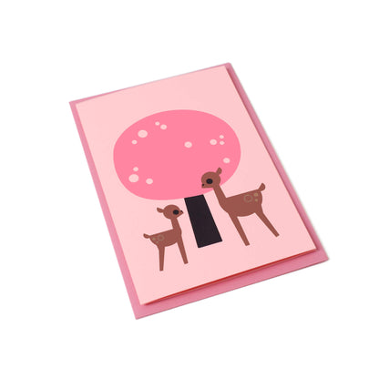 A tilted pink greeting card with an illustration of a cherry blossom tree and two deer, one small and one larger. A pink envelope is visible behind the card.
