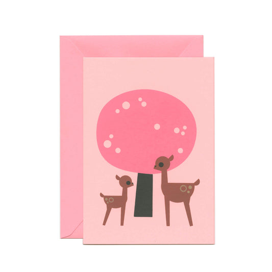 A pink greeting card with an illustration of a large and small deer standing in front of a pink tree. There's a pink envelope behind the card.