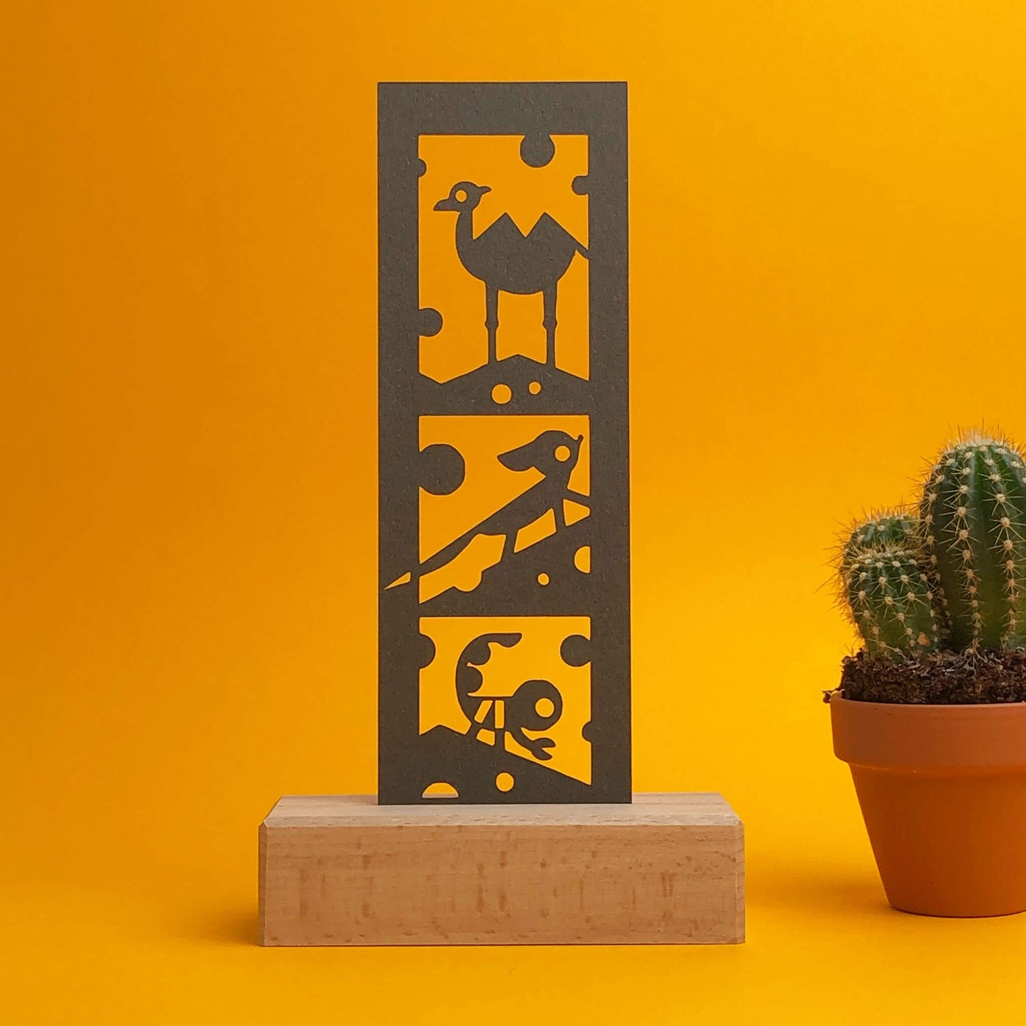  Wooden art display with paper bookmark cutouts of camel, fennec fox, and scorpion, accompanied by a small cactus in a terra cotta pot. Against an orange and yellow background.