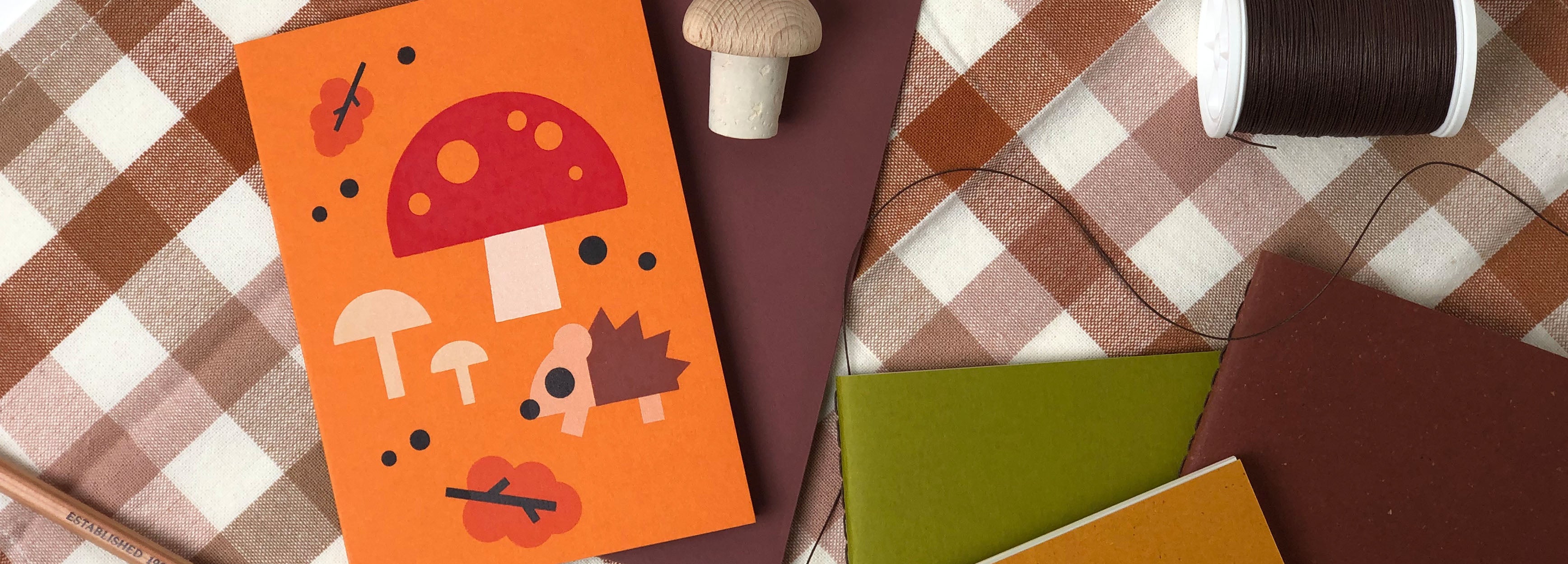 Cropped arrangement of three notebooks ranging from colors red-brown, green and yellow, an orange greeting card with illustrated hedgehog, mushrooms and leaves on a checkered cloth. Scattered are a decorative wooden mushroom, pencil and thread.