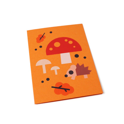 A tilted orange greeting card with an illustration of a hedgehog, mushrooms, leaves and scattered dots on a white background.