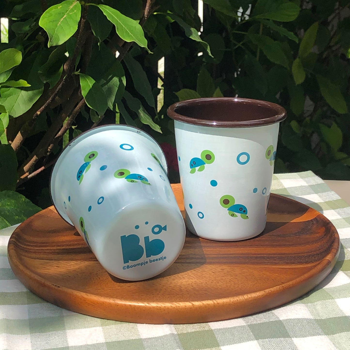 Pair of light blue cups with sea turtle design sits on a round wooden platter on a gingham green cloth. Surrounded by green leaves in the background.