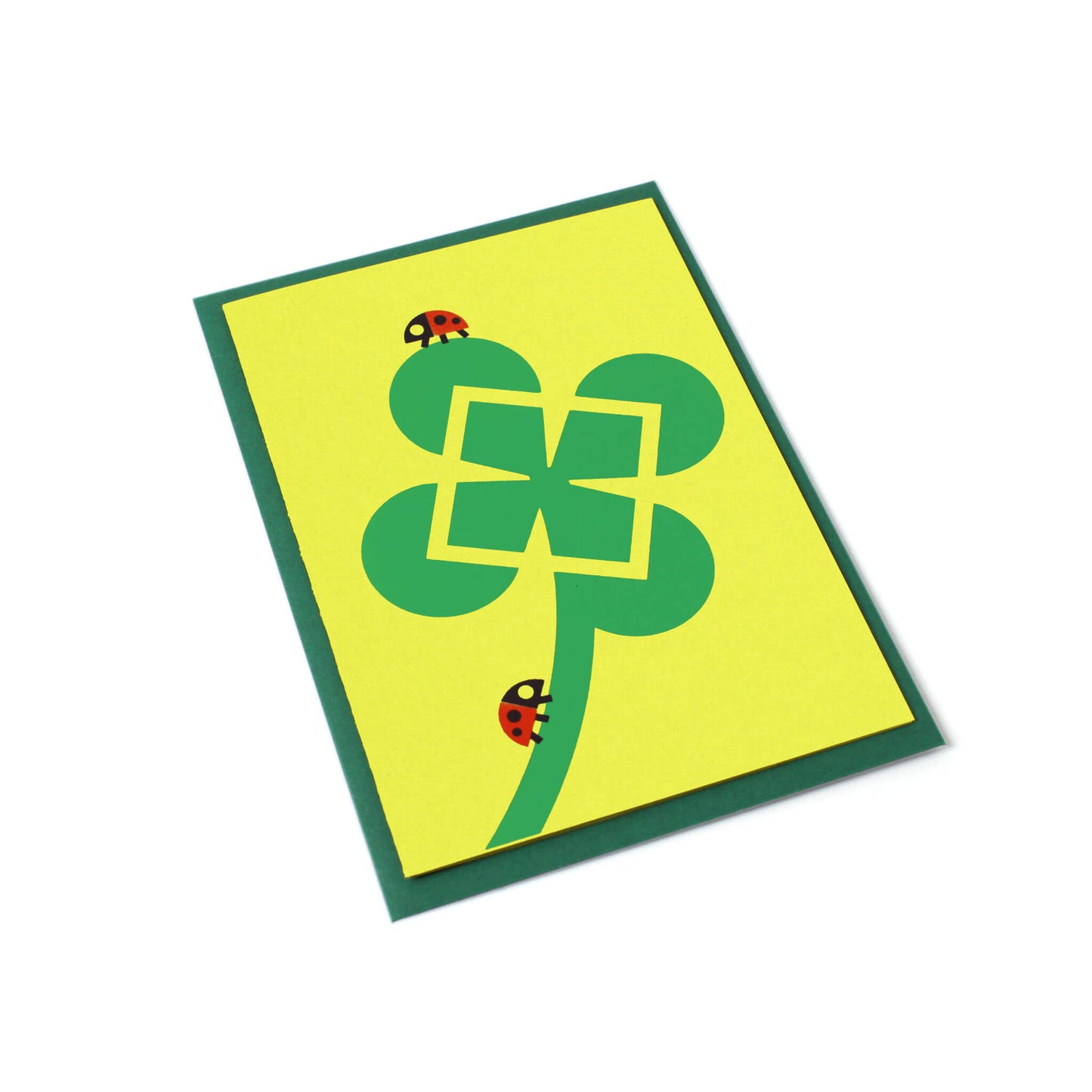 A tilted green greeting card with an illustration of a four-leaf clover and two ladybugs. Behind the card, there's a dark green envelope.
