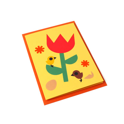 A yellow greeting card with a tulip, chicks, flowers, and egg illustration stands tilted, with an orange envelope behind it.
