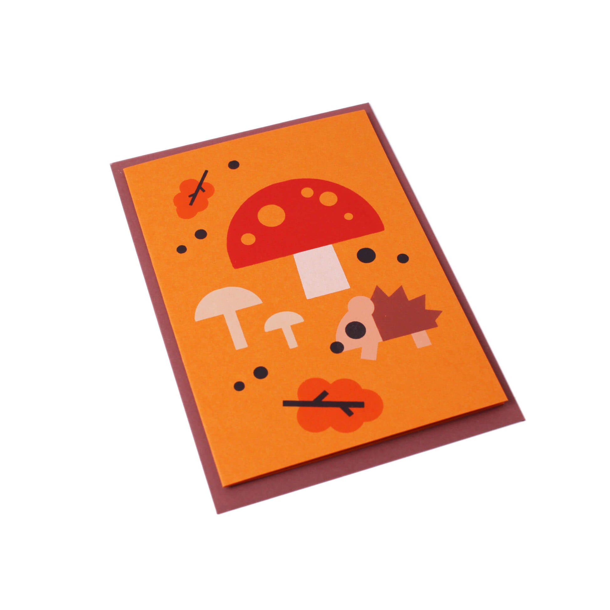A tilted orange greeting card with an illustration of a hedgehog, mushrooms, leaves and scattered dots in the background. A brown envelope is visible behind the card.