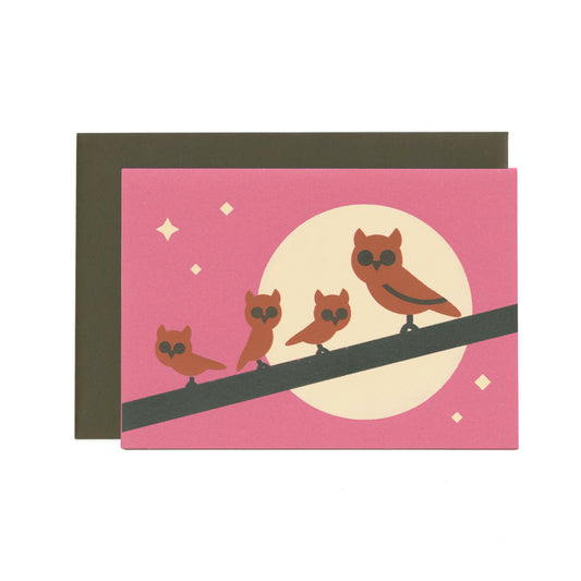 Purple greeting card featuring a family of brown owls on a branch with a moon and stars, and a dark brown envelope in the background.