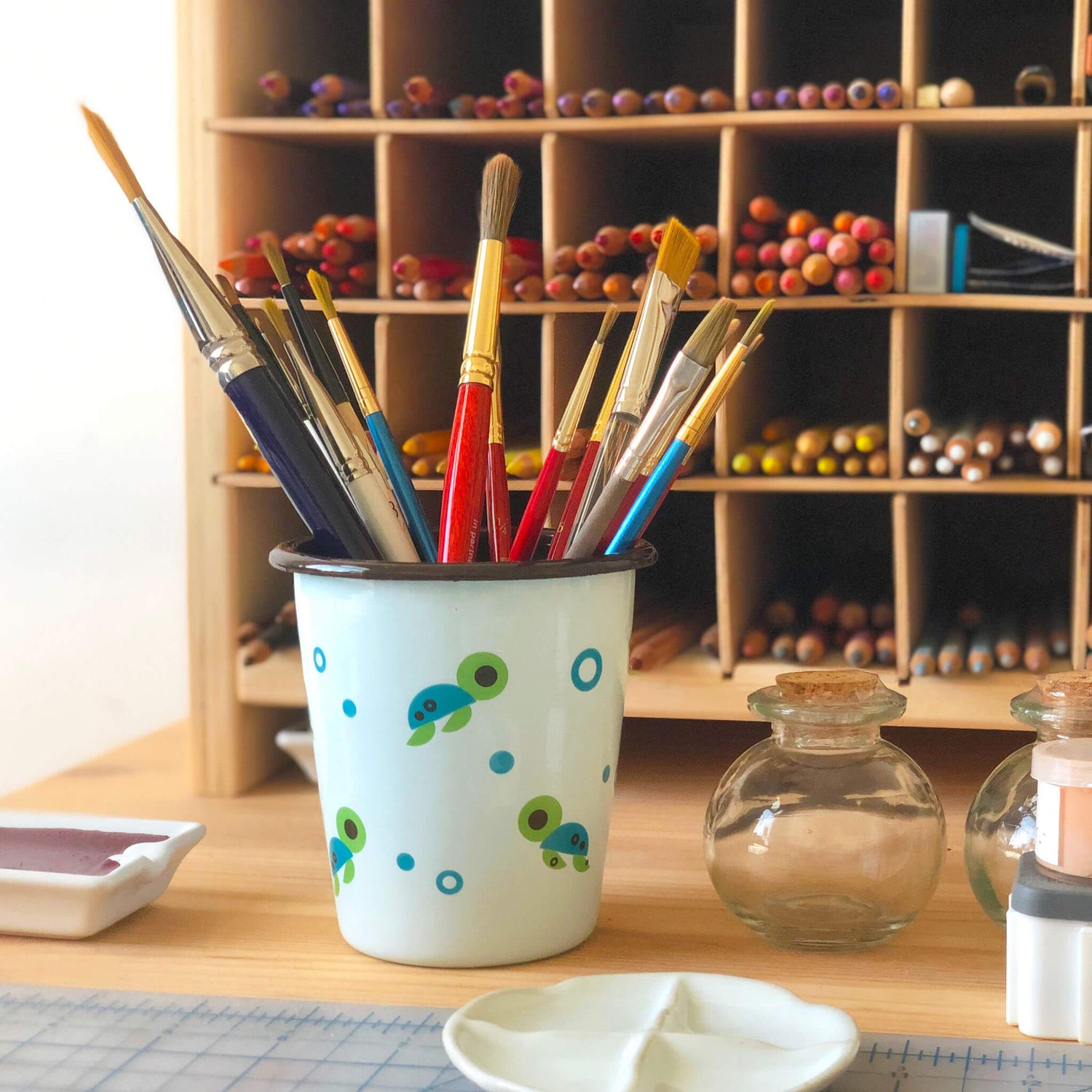 Assorted paintbrushes are held in a light blue pen cup decorated with a sea turtle illustration pattern on a wooden desk. Ceramic and glass containers are arranged nearby, and a storage holder contains colored pencils in the background.