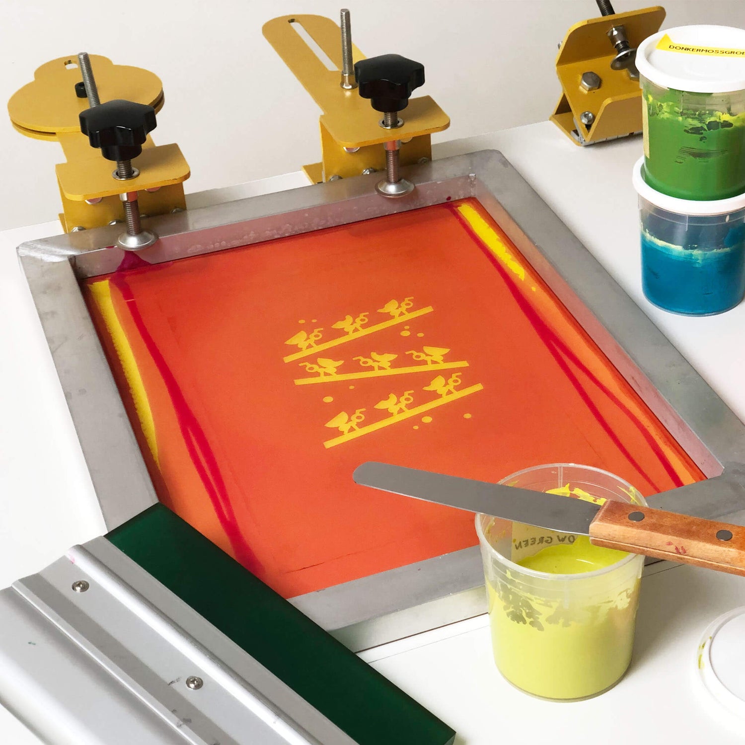 Screenprint supplies arranged on a table: a framed screen, a squeegee,  and three containers of three colored inks (green, blue, and yellow)  alongside. An open yellow ink container has a spatula on top.