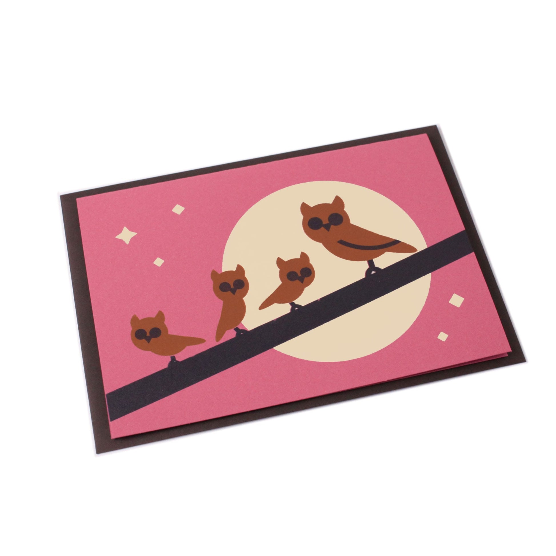 A tilted magenta-purple greeting card with an illustration of a branch with one big and three small brown owls and a moon with stars in the background. Behind the card is a dark brown envelope.