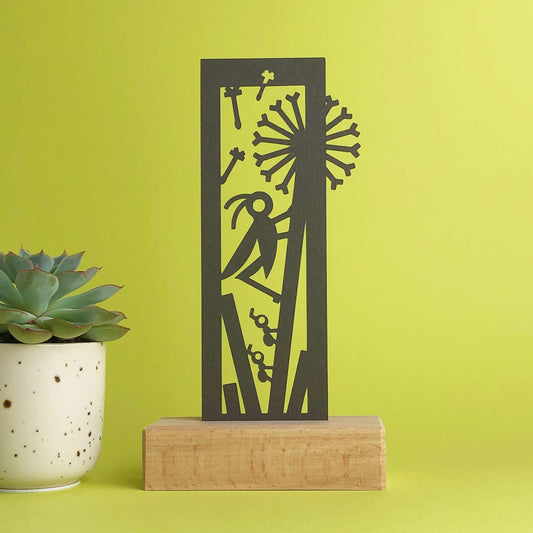 A  wooden art display showcasing a brown bookmark with cut paper art of a  dandelion and insects. Beside the display, a potted succulent sits  against a light green background.