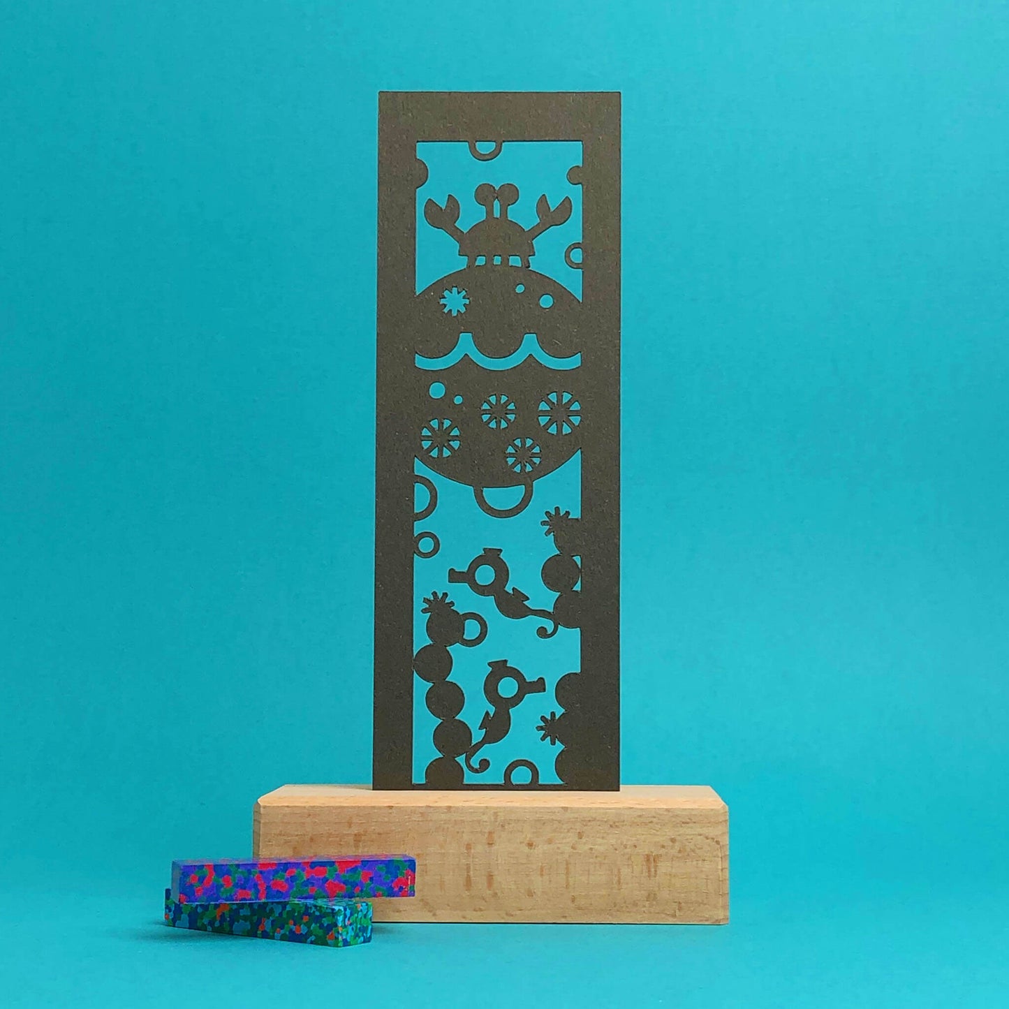A wooden art display with a brown paper bookmark featuring a cut-out designs of a crab, seahorses, and coral. Two speckled crayons, in shades of blue, green, and red, are placed beside the display against a blue background.