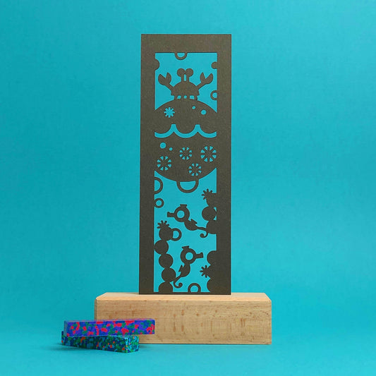 A wooden art display with a brown paper bookmark featuring a cut-out designs of a crab, seahorses, and coral. Two speckled crayons, in shades of blue, green, and red, are placed beside the display against a blue background.