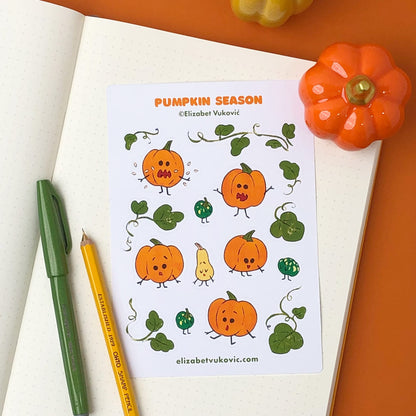 Pumpkins-themed sticker sheet sits on an opened notebook page. Beside it a pen, pencil and ceramic pumpkin are placed.